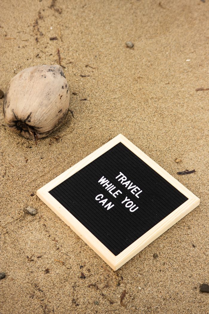 Inspirational travel message on a letter board placed on sandy beach with a coconut nearby.