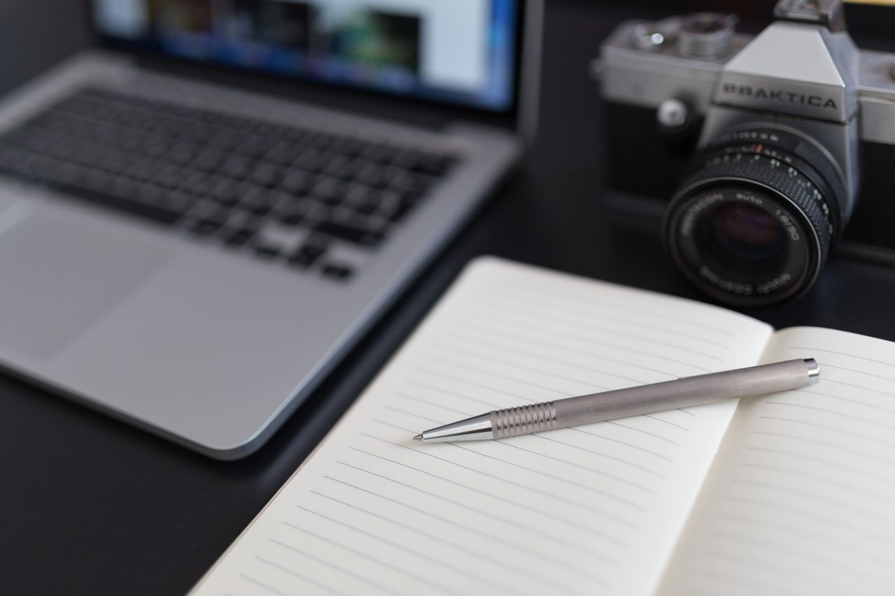 Offerings A close-up view of a laptop, vintage camera, and open notebook with a pen.